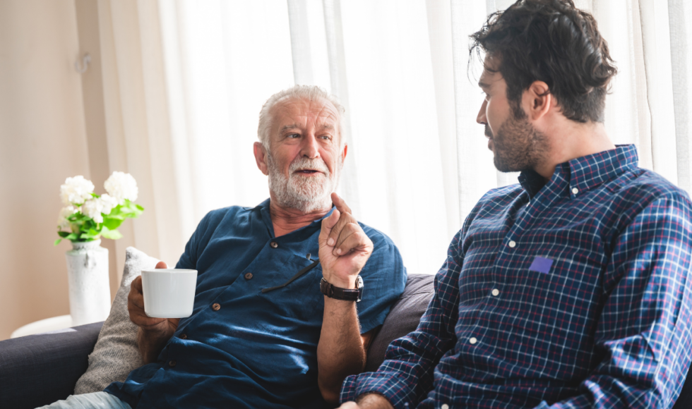 Older father and adult son sitting together on a couch having a meaningful conversation, illustrating family discussions about aging parent care, safety planning, and maintaining independence for seniors.