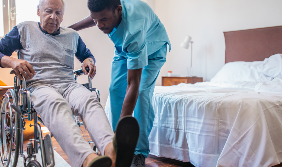 Older adult being assisted into a wheelchair by a caregiver in a nursing home bedroom, highlighting senior care support, mobility assistance, and risks of understaffing or neglect in long-term care facilities.