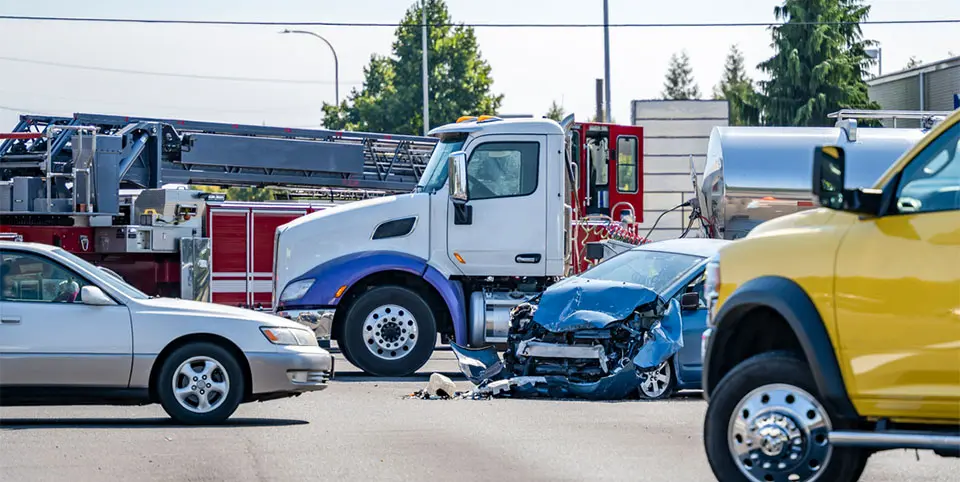 Severely damaged blue car at the scene of a collision involving a large commercial truck